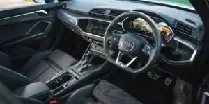 The image shows the interior of an Audi Q3 Sportback. The interior is mostly black with red accents. The car has a digital instrument cluster and a large central touchscreen. The seats are sporty and supportive, with a black and gray fabric pattern. The steering wheel is wrapped in leather and has red stitching.