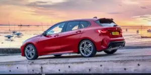 A red BMW 1 Series car is parked on a coastal road with a scenic sunset in the background. The car has a sporty design with large alloy wheels and a rear spoiler.