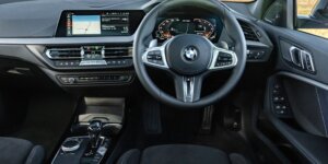 The interior of a BMW 1 Series car. The dashboard features a large touchscreen display, and the seats are upholstered in black fabric. The steering wheel has multiple buttons for controls, and the center console includes a gear selector and other buttons.