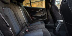 The rear seats of a BMW 1 Series car. The seats are upholstered in a combination of black fabric and Alcantara, and they have sport-style bolsters for added support. The rear door panels feature a simple design with a door pocket and a window control.