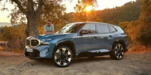 A man standing next to a blue BMW XM electric SUV. The car is parked in a scenic landscape with trees and a sunset in the background. The car has a futuristic design with large alloy wheels and a closed grille.