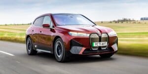 A red BMW iX electric SUV driving on a road with a scenic landscape in the background. The car has a futuristic design with large alloy wheels and a closed grille.