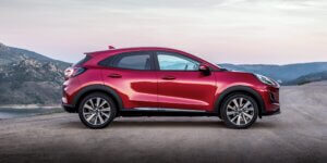 The image shows a side profile of a red Ford Puma compact SUV parked on a dirt road in a mountainous landscape. The car's bold red color contrasts sharply with the natural background of rugged hills and a distant body of water, under a soft, pastel-colored sky. The vehicle's design emphasizes its sporty and dynamic character, with a rising shoulder line, black wheel arches, and large alloy wheels. The overall setting suggests a sense of adventure and capability, highlighting the Puma's versatility in both urban and outdoor environments.