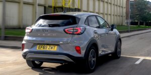 The image depicts the rear view of a silver Ford Puma compact SUV driving down a street in an urban setting. The car's distinctive design features include sleek, boomerang-shaped LED taillights and a subtle rear spoiler integrated into the roofline. The model name "Puma" is prominently displayed across the tailgate in chrome lettering. The rear license plate, reading "EF69 ZMT," is positioned centrally on the bumper. The vehicle's smooth curves and modern aesthetics are highlighted as it drives past a large industrial or commercial building with vertical paneling and a green lawn area.