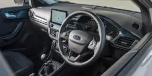 Interior view of a car's dashboard and steering wheel. The dashboard features a central touchscreen display, various control buttons, and air vents. The steering wheel, bearing the Ford logo, has built-in controls. The instrument panel behind the steering wheel shows gauges and meters. The driver's side door is visible with a door handle and control buttons.