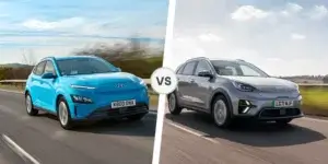 A side-by-side comparison of the Hyundai Kona Electric and Kia e-Niro. The Hyundai Kona Electric is blue and driving on the left side of the road. The Kia e-Niro is silver and driving on the right side of the road. Both cars are driving on a road with green fields in the background.