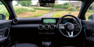 The interior of a Mercedes-Benz A-Class car. The dashboard features a large touchscreen display, and the seats are upholstered in black leather. The steering wheel has multiple buttons for controls, and the center console includes a gear selector and other buttons.