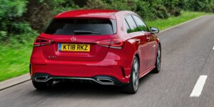 A red Mercedes-Benz A-Class hatchback car driving down a country road. The car is in motion, and the background is blurred due to the speed. The rear of the car is visible, showing the taillights, rear bumper, and license plate.