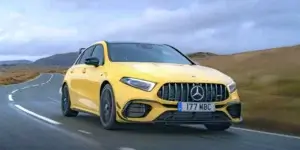 Front view of a Mercedes-Benz A 45 S, a high-performance hatchback, driving on a road. The car features a sleek, aerodynamic design with a prominent grille and LED headlights. The car is painted in a vibrant color, and the road and scenery in the background are slightly blurred, emphasizing the vehicle's speed.