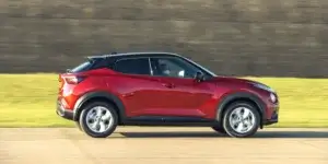 A red Nissan Juke driving down a road with a grassy field in the background. The car has a sporty design with black accents and alloy wheels.