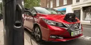 A red Nissan Leaf electric car is parked on a city street and plugged into a charging station. A London taxi is visible in the background.