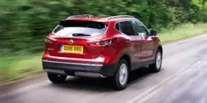 A red Nissan Qashqai driving on a winding road with trees on either side. The rear of the car is visible, showing the taillights, rear spoiler, and alloy wheels.