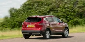 A red Nissan Qashqai driving on a winding road with trees on either side. The rear of the car is visible, showing the taillights, rear spoiler, and alloy wheels.