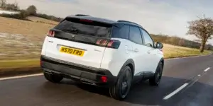 A white Peugeot 3008 SUV driving down a winding road with a rural landscape in the background. The rear of the car is visible, showing the taillights, rear spoiler, and license plate.