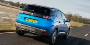 A blue Peugeot 3008 SUV driving down a road with a rural landscape in the background. The rear of the car is visible, showing the taillights, rear spoiler, and license plate.