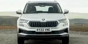 A white Skoda Karoq SUV parked on a dirt road with a scenic mountain backdrop. The front of the car is visible, showing the headlights, grille, and license plate "KY22 VNE".