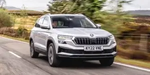 A white Skoda Karoq SUV driving down a winding road in a rural landscape. The car has a modern and sleek design with black accents and alloy wheels. The license plate reads "KY22 VNE".