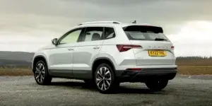 A white Skoda Karoq SUV parked on a dirt road with a scenic mountain backdrop. The rear of the car is visible, showing the taillights, rear spoiler, roof rack, and black alloy wheels. The license plate reads "KY22 VNE"