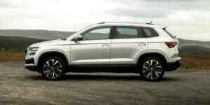 A silver Skoda Karoq SUV parked on a dirt road with a scenic mountain backdrop. The car has black alloy wheels, a roof rack, and a modern, sleek design.