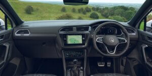 The interior of a Volkswagen Tiguan. The car features a black dashboard with a large central touchscreen display, a leather-wrapped steering wheel, and comfortable-looking front seats. The view outside the windshield shows a rural landscape with rolling hills and green fields.