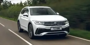 A white Volkswagen Tiguan R-Line SUV driving down a winding road in a rural landscape. The car has a sporty design with black accents and alloy wheels. The license plate reads "T2 VWN".