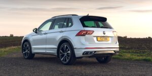 A silver Volkswagen Tiguan R-Line SUV parked on a dirt road with a field of grass in the background. The rear of the car is visible, showing the taillights, rear spoiler, and black alloy wheels. The license plate reads "T2 VYB"