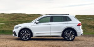 A white Volkswagen Tiguan R-Line SUV parked on a dirt road with a scenic mountain backdrop. The car has black alloy wheels, a roof rack, and a modern, sleek design.