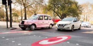 A pink London taxi and a white Nissan Leaf electric car driving on a city street. The black circle of a congestion charge zone is painted on the road.