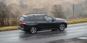 A black BMW X7 driving on a wet road surrounded by green hills. The car has a roof rack and is seen from the side. There are clouds in the sky, suggesting it's a cloudy day.