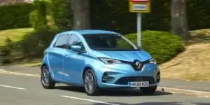 A blue Renault Zoe electric car is driving on a road with trees and a street sign in the background. The car has alloy wheels and is moving at a moderate speed.