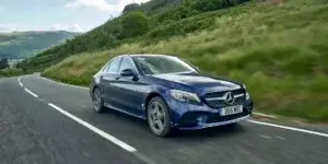 A blue Mercedes-Benz drives on a winding road through a lush, green landscape with hills in the background.