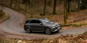 A gray Mercedes-Benz GLE SUV driving on a winding road through a forest. The SUV has black alloy wheels and a roof rack.