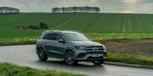 A gray Mercedes-Benz GLS SUV driving on a winding road with a rural landscape in the background.