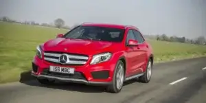 The car in the image is a red Mercedes-Benz GLA. It appears to be driving on a country road. The car has a sporty design with a large front grille and headlights. It is also equipped with a sunroof.