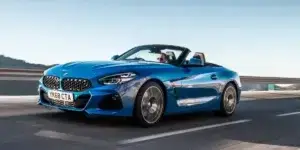 A blue BMW Z4 Roadster driving on a winding coastal road with a scenic backdrop. The top is down and the driver is visible.