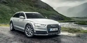 A silver Audi A6 Allroad driving on a winding road surrounded by mountains. The car is facing the camera and has a rugged appearance with black plastic cladding around the wheel arches and side skirts. The license plate on the car reads "JF2 323.