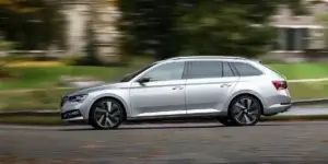 A silver Skoda Superb Combi estate car driving on a road with trees and houses in the background. The car is in motion, and the background is blurred due to the speed.