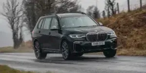 A black BMW X7 SUV driving on a wet road with a scenic landscape in the background. The car has a large size, a sporty design with a sloping roofline, and large alloy wheels.