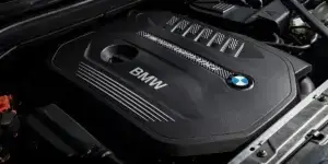 A close-up of a BMW engine block with the BMW logo prominently displayed. The engine cover is black and has a sleek design.