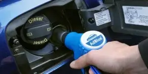 Image showing a person holding a blue plastic container labeled 'AdBlue' near a car's fuel cap. The car's fuel filler area is open, ready to receive the AdBlue fluid, which is used to reduce emissions in diesel engines.