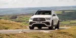 The car in the image is a white Mercedes-Benz GLE. It appears to be driving on a winding road in a rural area. The car has a sporty design with a large front grille and headlights. It is also equipped with a sunroof.