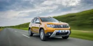 A yellow Dacia Duster SUV driving on a winding road surrounded by green fields. The sky is cloudy, and the car is moving at a high speed.