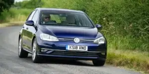 A blue Volkswagen Golf parked on a roadside with greenery in the background.