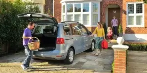 A family of four standing in front of a silver minivan in their driveway. The minivan's trunk is open, and a man is loading a picnic basket into it. The woman is holding a red beach ball, and the two children are standing next to her.