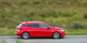 A red Opel Astra hatchback car is driving on a winding road surrounded by green hills. The car has alloy wheels and appears to be moving at a moderate speed.