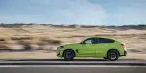 A green BMW X4 M driving on a road with a desert landscape in the background. The car is in motion, and the background is blurred due to the speed.