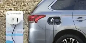 A silver Mitsubishi Outlander PHEV is plugged into a charging station. The car is parked in a driveway with a stone wall in the background. The charging station has a white cable and a display screen showing charging information.