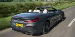 A gray BMW 8 Series Convertible driving on a winding road with a rural landscape in the background. The top is down.