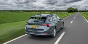 A gray Skoda Octavia Combi estate car driving on a road with a field and a clear sky in the background. The car is in motion, and the rear of the car is visible, showing the taillights, rear bumper, and license plate.