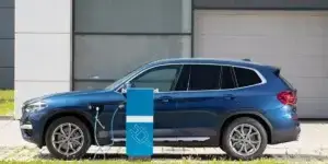 A blue BMW X3 plug-in hybrid car parked next to a charging station. The car has a charging cable plugged into the charging port on the front fender.